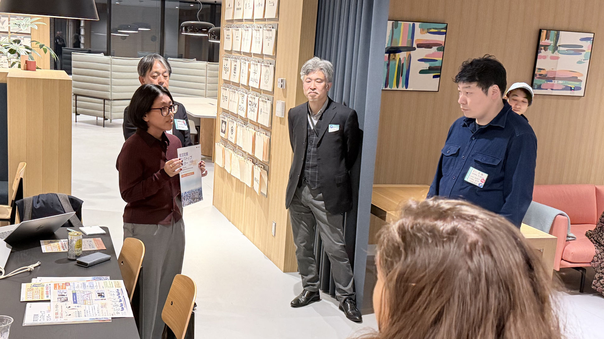 フリーランスMeetupの会場で、スタッフが資料を手に持って説明している様子。周囲には数名の参加者が立って話を聞いている。テーブルにはイベントの案内資料やPCが置かれている。